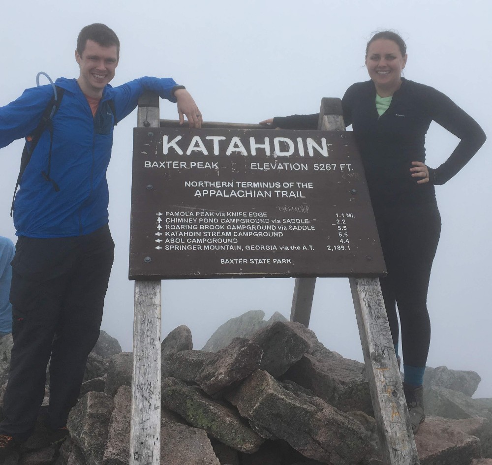 A man and a woman stand in the fog next to a sign marking Katahdin's peak